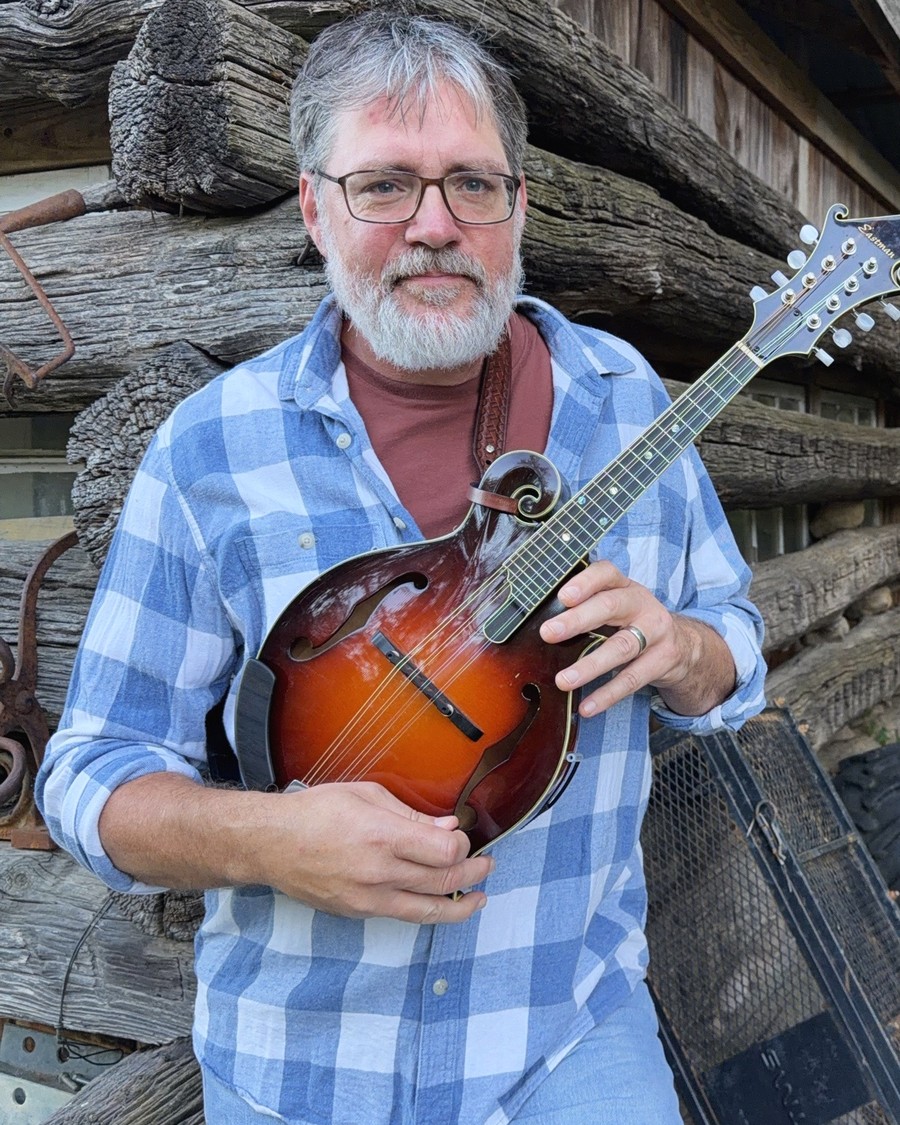 William leaning against a log outbuilding holding an Eastman F-style mandolin.