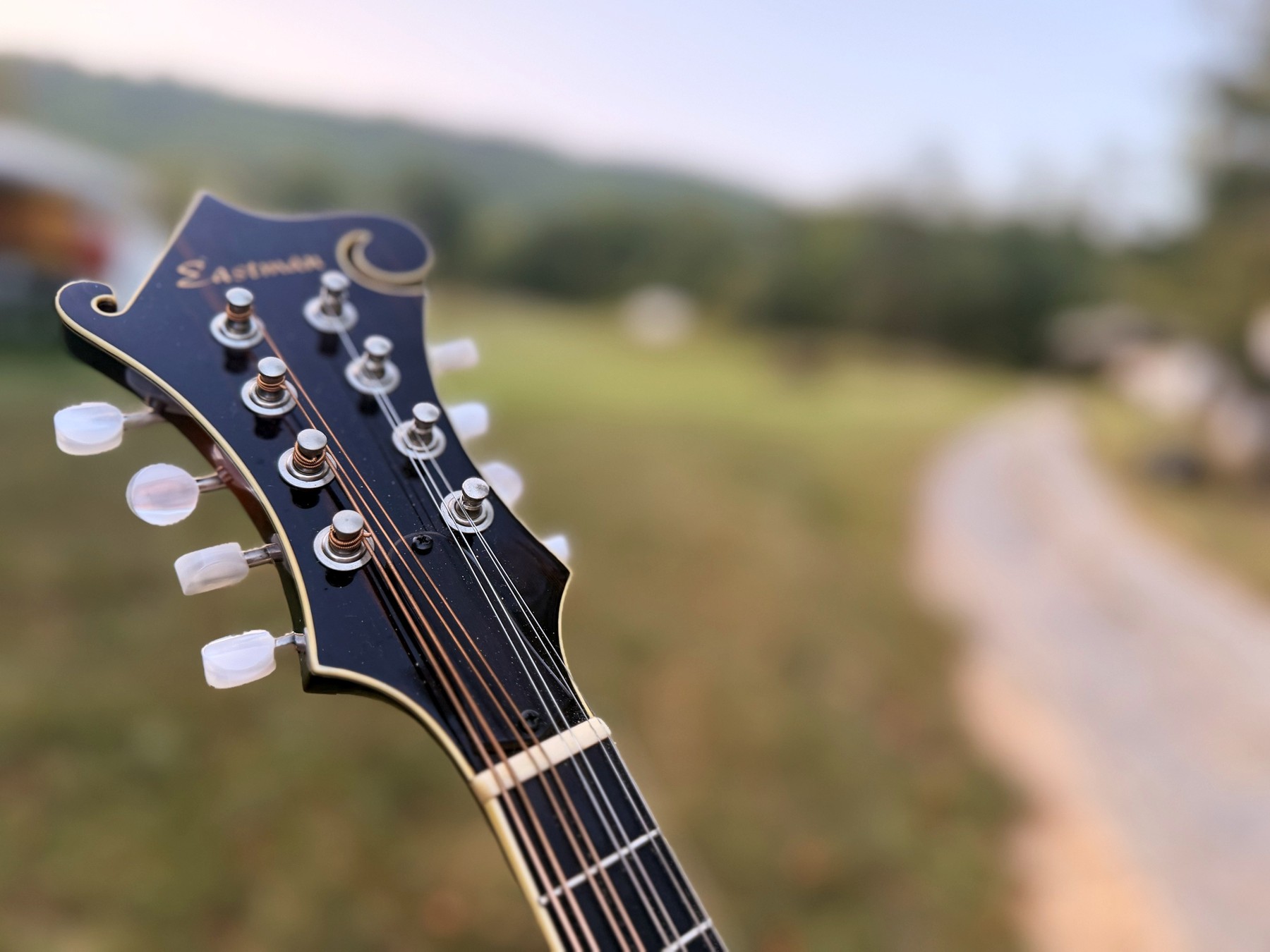 The Eastman mandolin's scrolled headstock and tuners in low evening light, a farm track falling away behind it.